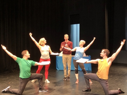 A man in a maroon shirt in the middle of four dancers, two on either side with their arms stretched upwards.