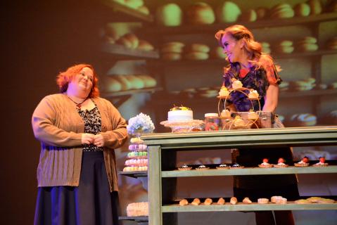 Genevieve and Hortense stand by the bakery counter. It features a colorful macaron tower, a cake, cupcakes displayed on a ferris wheel themed stand, fruit tarts, and more