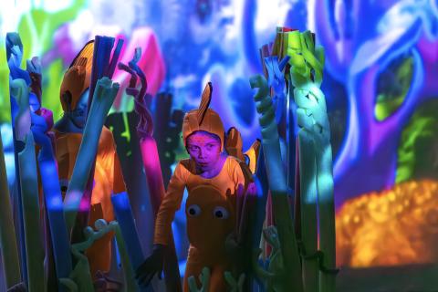 A young performer dressed in an orange fish costume peeking out through coral stage props against a digital backdrop of the sea and coral.