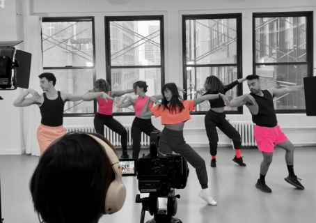 Five dancers wearing pink tops and shorts in a row demonstrating a dance move while someone is filming in front of them.