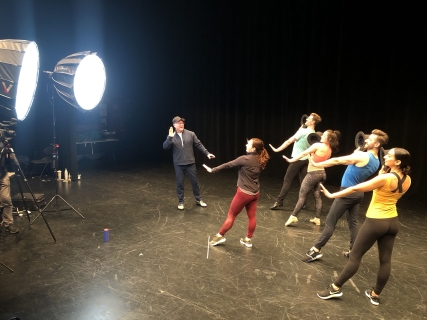 A choreographer demonstrating a move as four dancers behind perform the movement in front of bright lights.