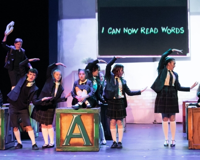 Young performers in school uniforms onstage with letter blocks and classroom projections