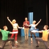 A man in a maroon shirt in the middle of four dancers, two on either side with their arms stretched upwards.