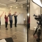 Four dancers stand in a row with their arms in their air and feet together in front of a filming camera.