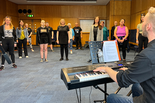 Teachers standing in a singing rehearsal with a man sat at the piano.